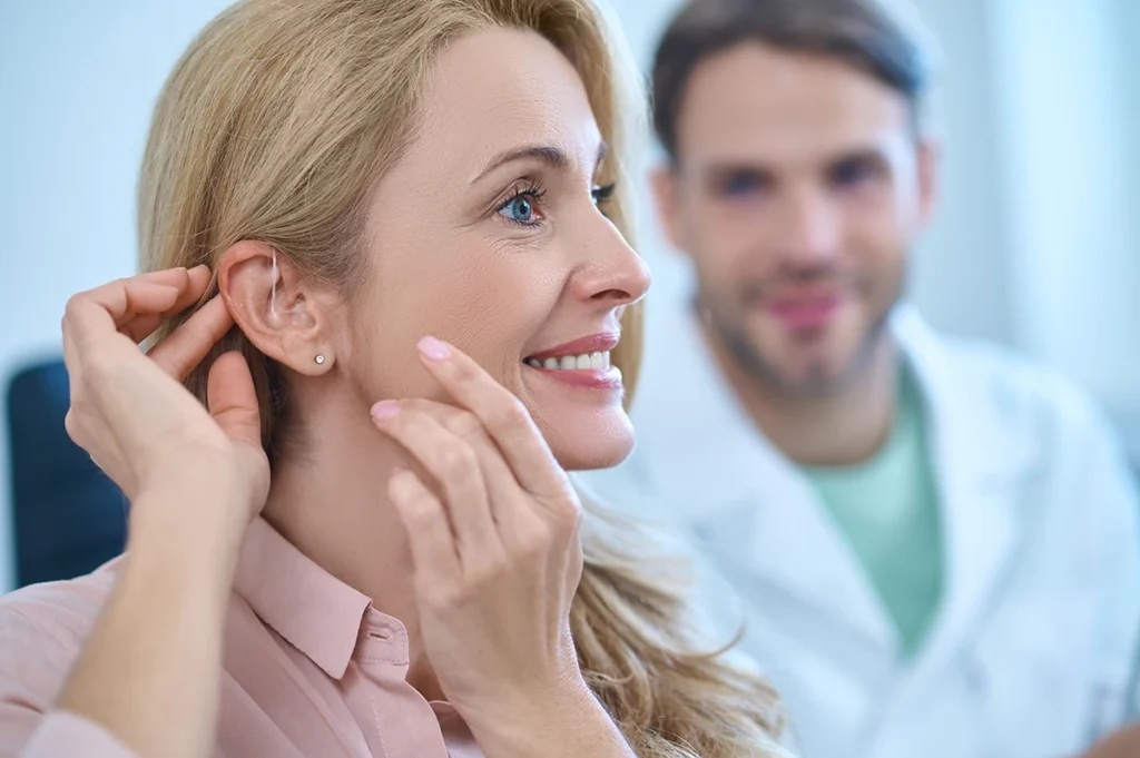 An older adult women smiling while putting in an AI hearing aid while at a hearing health care clinic.